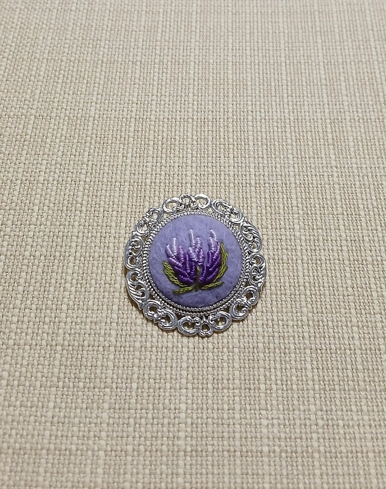 Silver ornate brooch with purple enameled center featuring a dark purple lotus flower design on textured beige fabric background