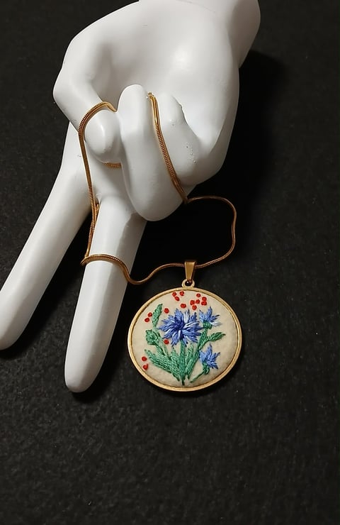 White ceramic hand wearing a delicate embroidered flower pendant necklace with blue and red floral design on dark background