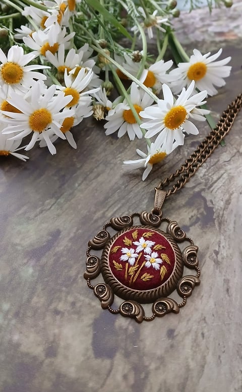 Ornate bronze pendant necklace with red enamel daisy design next to fresh white daisies on gray surface