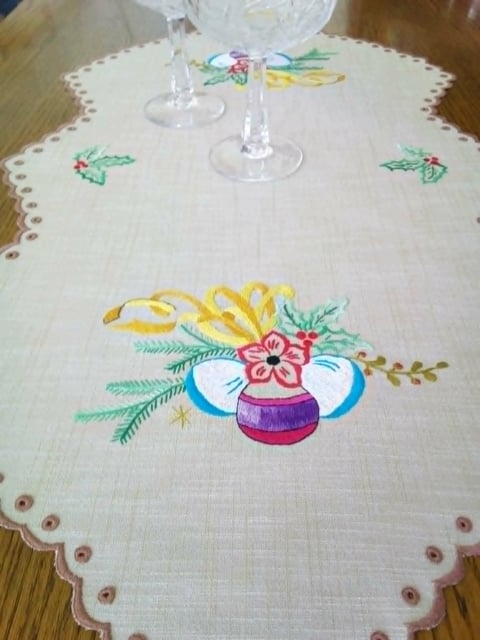 Embroidered cream table runner with festive holiday ornaments and floral designs, scalloped edges, displayed on wooden table with glasses
