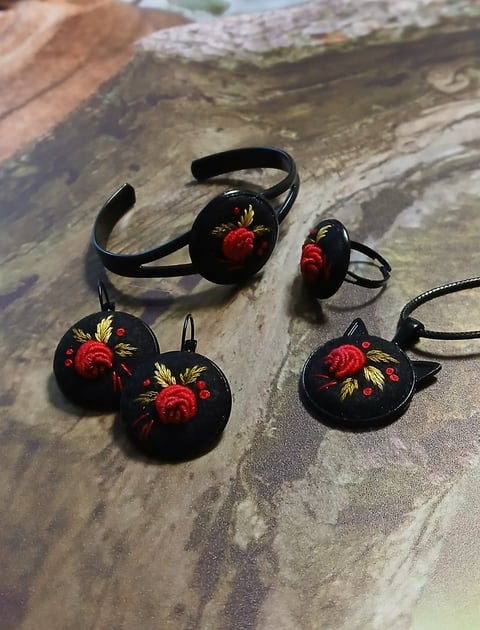 Black jewelry set with red rose embroidery designs including bangles, rings, and earrings on a textured wooden surface