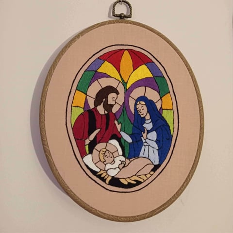 Oval embroidered nativity scene with Mary and Joseph in traditional robes around baby Jesus, set in wooden frame with stained glass style background