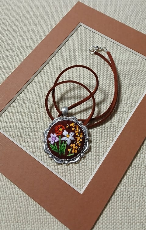 Embroidered floral pendant on burgundy cord necklace with scalloped metal frame on tan and brown matted background