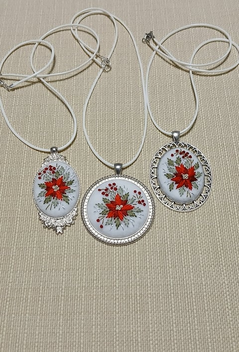 Three silver pendant necklaces with red floral designs on white backgrounds, displayed on a beige textured surface.