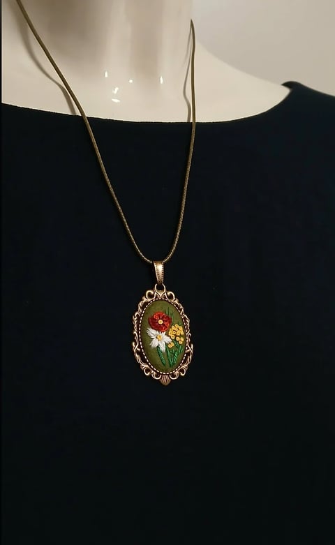 Gold pendant necklace with ornate oval frame containing red and white flowers on green background, worn on black garment