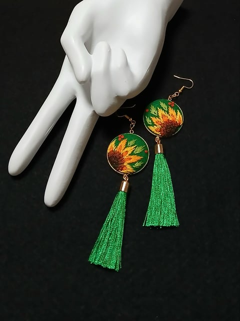 Pair of dangling earrings with sunflower design spheres and green tassel details displayed on white mannequin hand