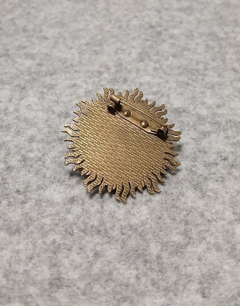Gold square brooch pin with textured surface and spiky fringe edges on gray background