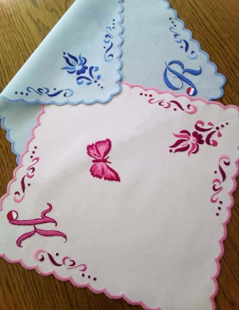 Two decorative embroidered handkerchiefs with scalloped edges, one white with blue designs and one white with pink floral embroidery
