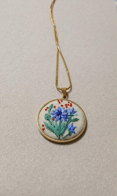 Gold pendant necklace with circular embroidered medallion featuring blue flowers, green leaves, and red berries on cream fabric background