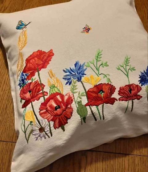 Cream-colored throw pillow with embroidered red poppies, blue cornflowers, wheat stalks, and green ferns on a wooden floor