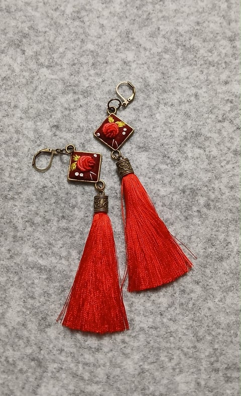 Two handmade key chains with red enamel square pendants featuring floral designs and matching red fabric tassels on a gray textured background