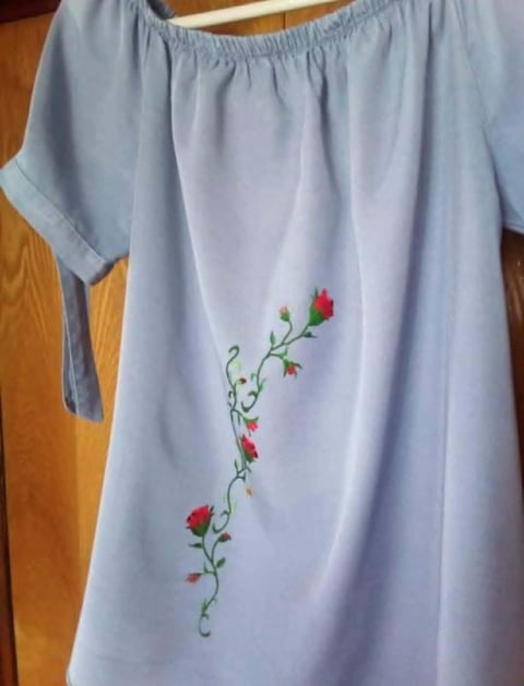 Light blue off-shoulder blouse with embroidered red and pink flowers trailing down the front, hanging on a wooden hanger