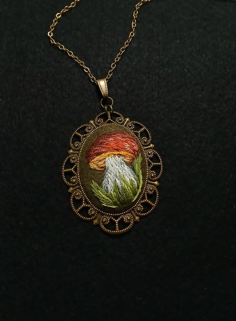 Gold filigree pendant necklace with ornate oval frame containing embroidered flower with red petals and green stem on dark background