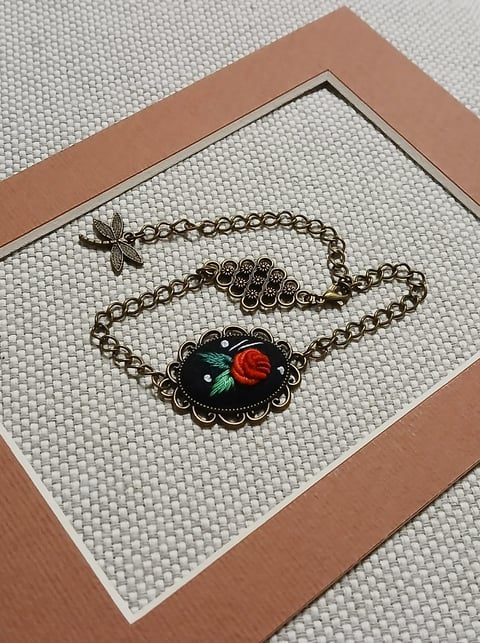 Ornate vintage-style bracelet with red rose and black cameo pendant on chain, displayed in tan frame on white textured background