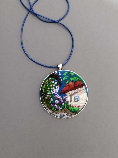 Pendant necklace with hand-painted circular design featuring a cottage, garden flowers, and blue sky on blue cord