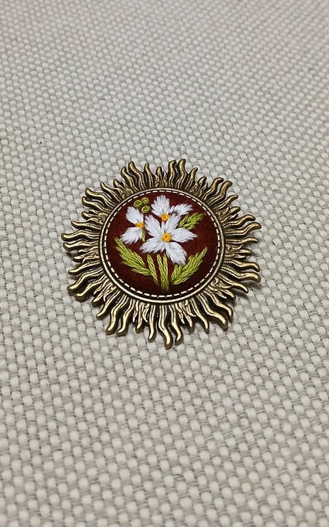 Decorative metal brooch with white daisy flowers and green leaves on dark red enamel background, ornate sun-like frame