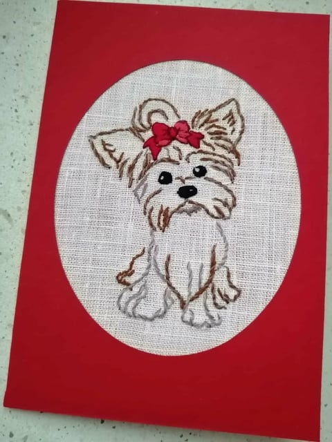 Embroidered illustration of a small dog with tan and white fur wearing a pink bow, centered on a red card