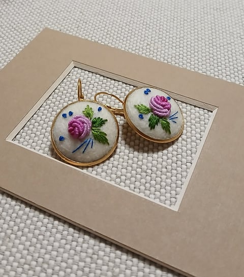 Pair of circular embroidered earrings with pink roses, green leaves, and blue flowers in brass settings on display