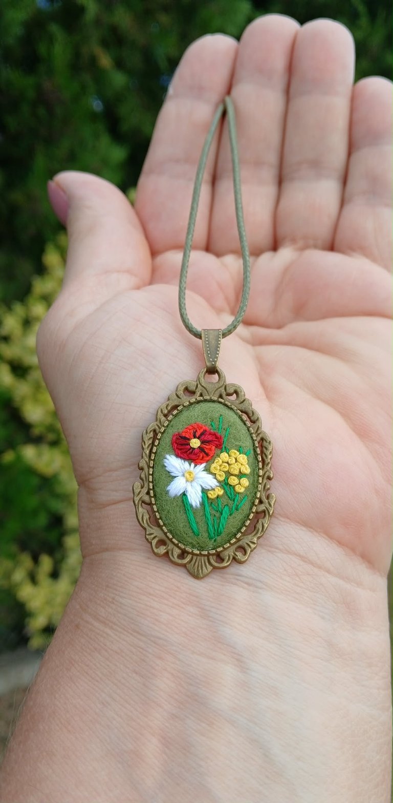 Hand holding embroidered floral pendant necklace with red, white, and yellow flowers on green fabric in ornate bronze frame