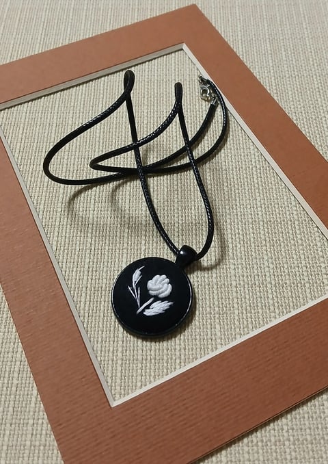 Black pendant necklace with white flower design on black circular charm, displayed on tan mat with brown frame