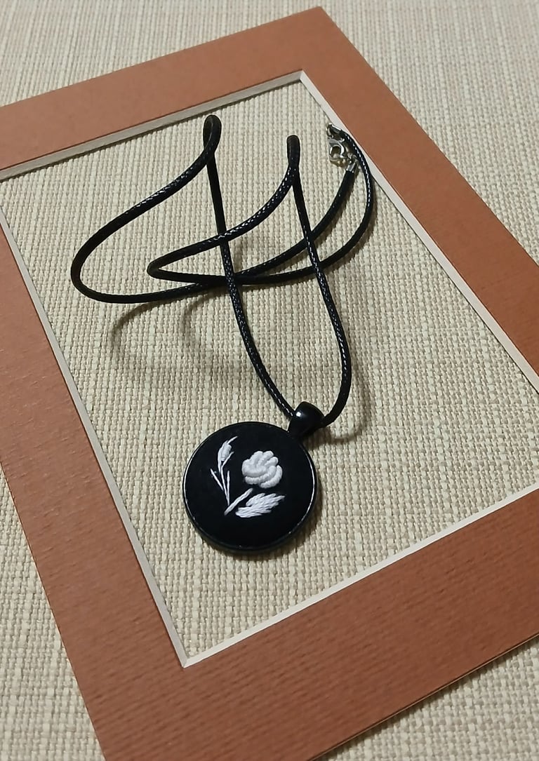 Black pendant necklace with white flower design on black circular charm, displayed on tan mat with brown frame