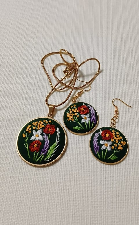 Embroidered floral jewelry set on dark green background featuring wildflowers in red, yellow, purple and white with gold accents.