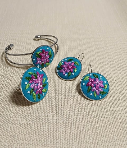 Set of blue enamel jewelry with pink floral designs, including a cuff bracelet and matching earrings on a neutral fabric background