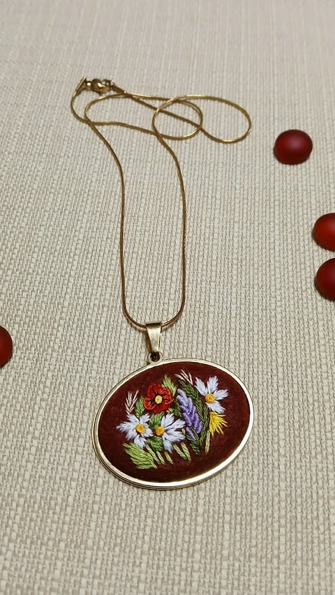 Gold chain necklace with oval pendant featuring colorful embroidered flowers on dark red background