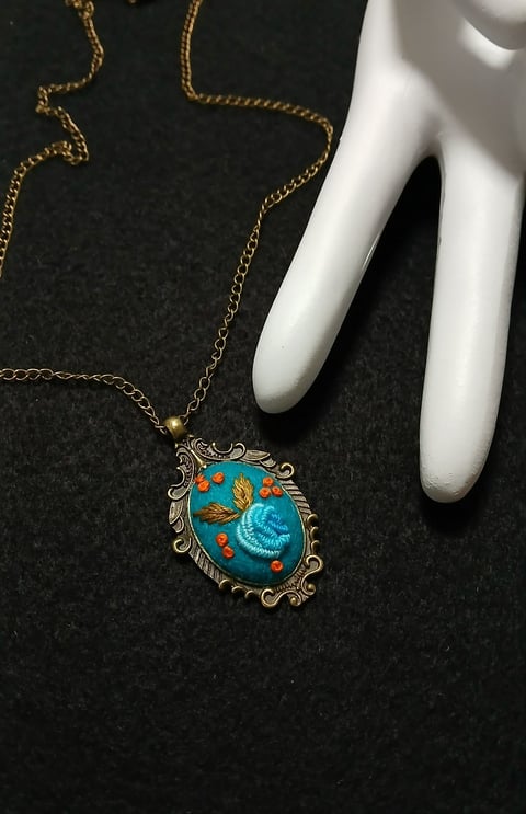 Gold chain necklace with ornate vintage pendant featuring blue enamel design and floral motifs, displayed on white mannequin hand against dark background