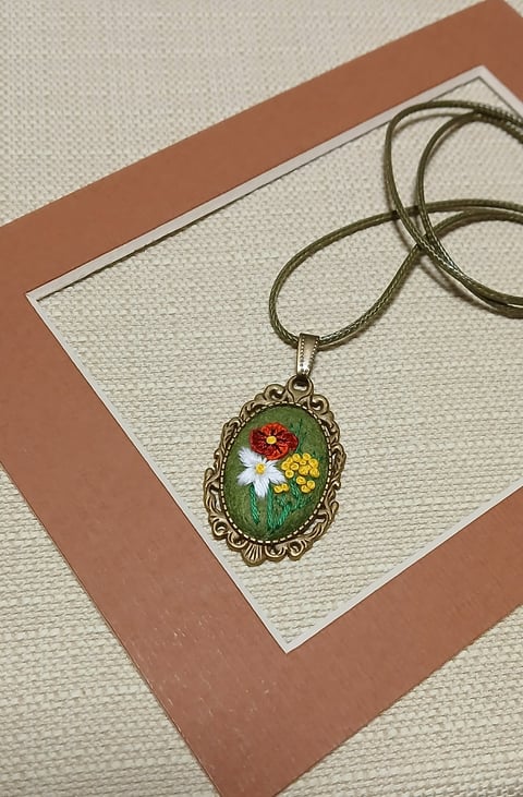 Ornate oval pendant necklace with colorful embroidered flowers on green background, displayed on brown and cream paper