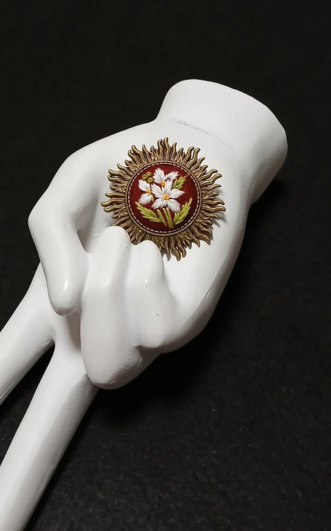 White mannequin hand holding ornate brooch with gold sunburst frame and white flowers on dark background