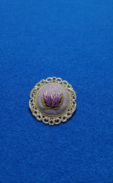 Ornate gold brooch with purple thistle flower on gray background against bright blue fabric