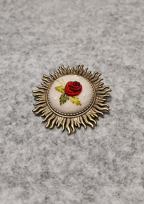 Ornate brass brooch with scalloped edge featuring a red rose on cream background against gray concrete surface