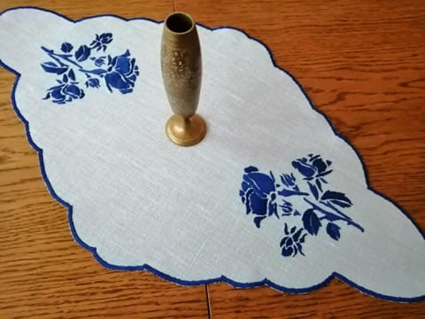 White linen table runner with scalloped edges and blue floral embroidery, topped with a brass flower holder.