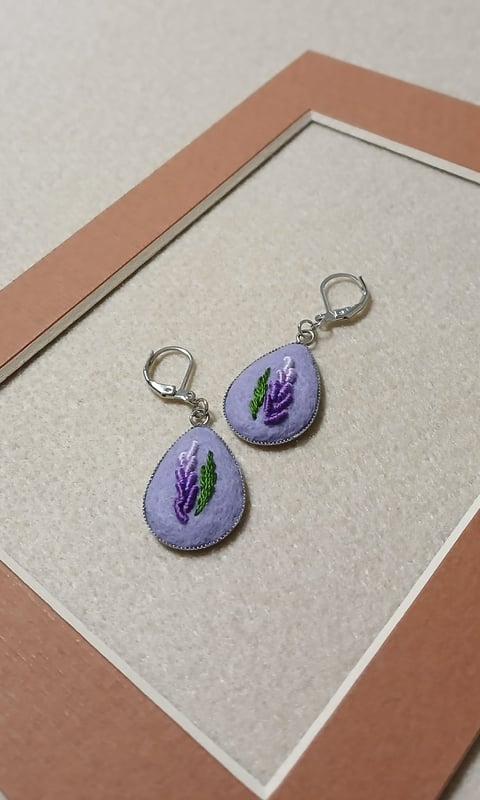 Pair of teardrop-shaped earrings with purple embroidered lavender flowers and silver metal clasps on a textured mat