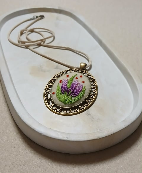 Hand-embroidered pendant necklace with purple flowers on ceramic dish, featuring ornate metal frame and beige cord