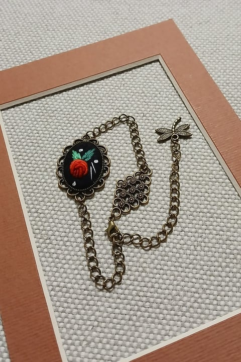 Ornate bronze chain bracelet with red rose pendant and dragonfly charm on white display mat with peach frame