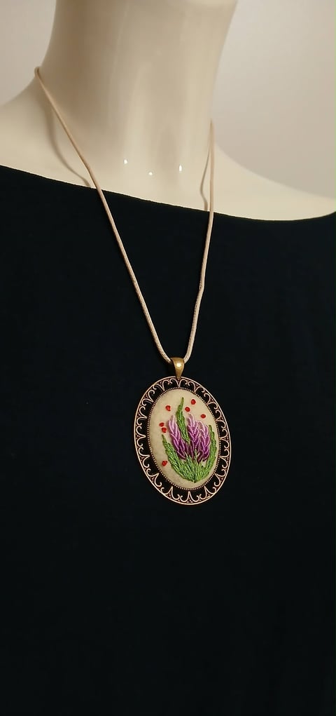 Embroidered floral pendant necklace with pink and purple flowers on cream fabric, hanging from a thin metallic chain