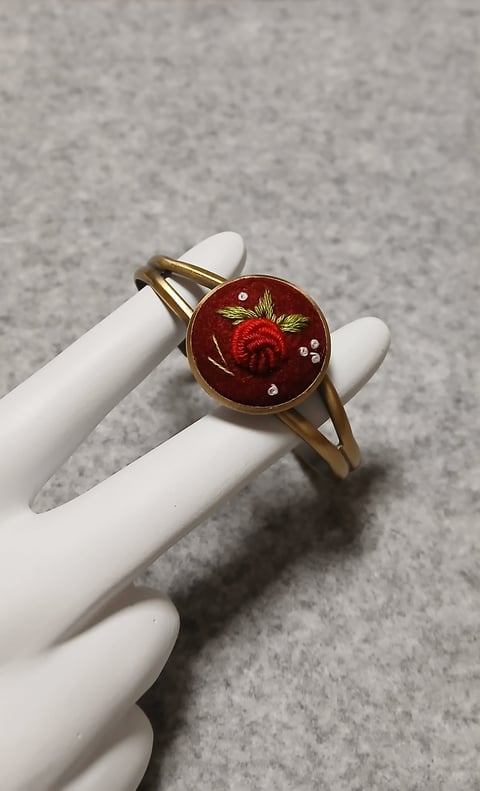 Gold ring with red enamel face featuring embroidered red rose and rhinestones, displayed on white mannequin hand
