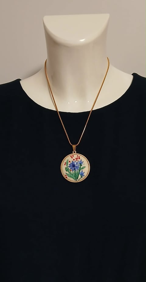 Mannequin wearing a black top and gold snake chain necklace with a round embroidered flower pendant featuring blue and red botanical design