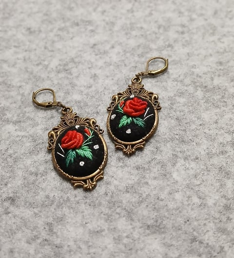 Pair of ornate bronze-framed earrings with black enamel backgrounds and red rose designs with green leaves on a gray surface