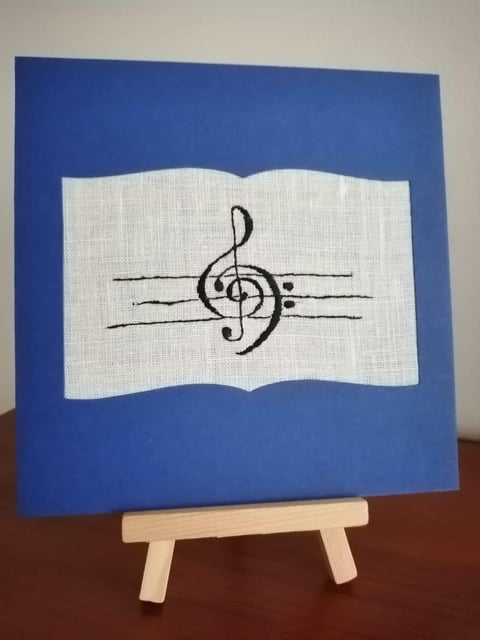 Handmade greeting card with treble clef musical note on white fabric patch against blue background, displayed on wooden easel