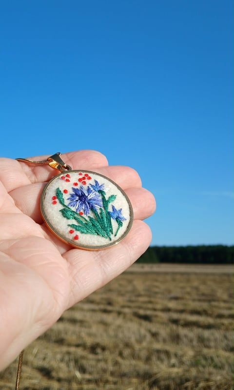 Hand holding embroidered pendant with blue flower and colorful details against outdoor sky and field background