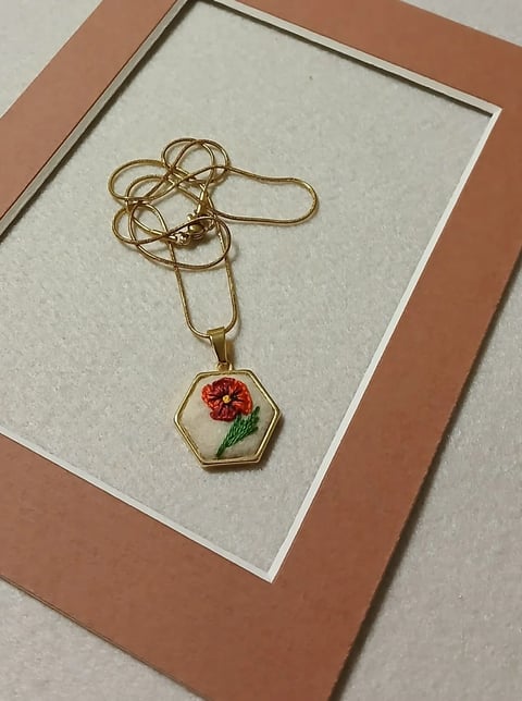Gold chain necklace with hexagonal pendant featuring embroidered red flower and green leaf on cream background