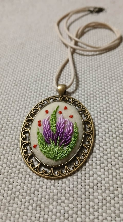 Ornate brass pendant necklace with embroidered purple flowers and green stems on white background