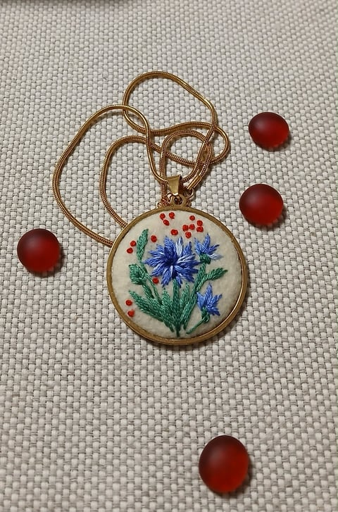 Embroidered pendant necklace with blue flowers and red berries in brass setting, with four red beads arranged around it on white fabric
