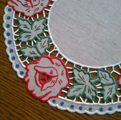 Decorative fabric doily with red rose designs, green leaves, white lace edging with blue beads, and plain linen center on wooden surface