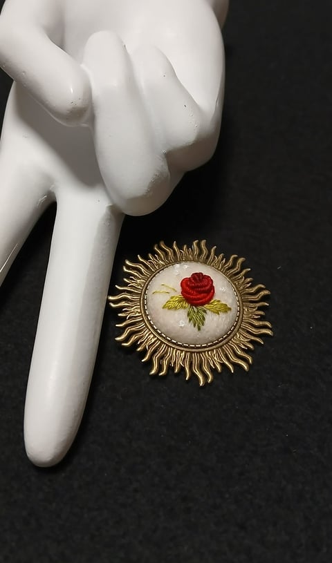 White porcelain hand figurine next to ornate gold-framed brooch with red rose embroidery on black surface