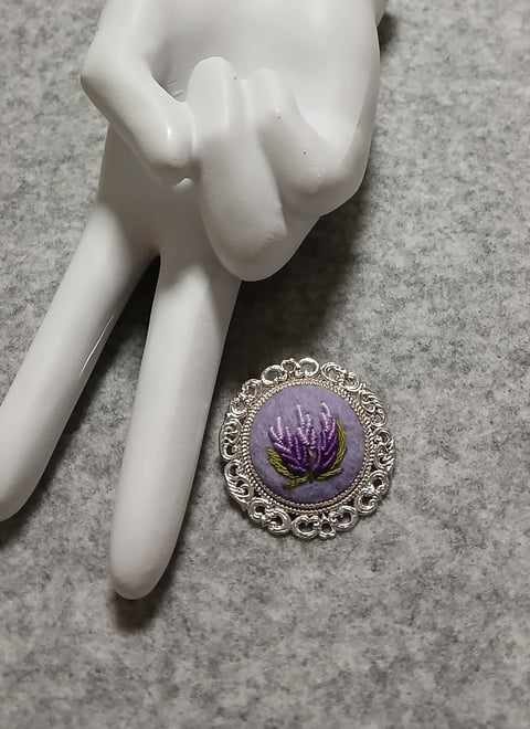 Silver ornate brooch with purple pressed flower on gray background next to white mannequin hand