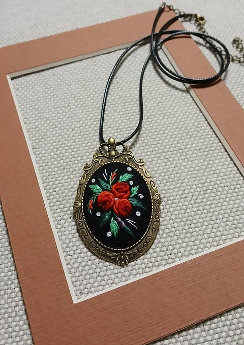Ornate oval pendant necklace with red rose design on black background, displayed on a tan and white card